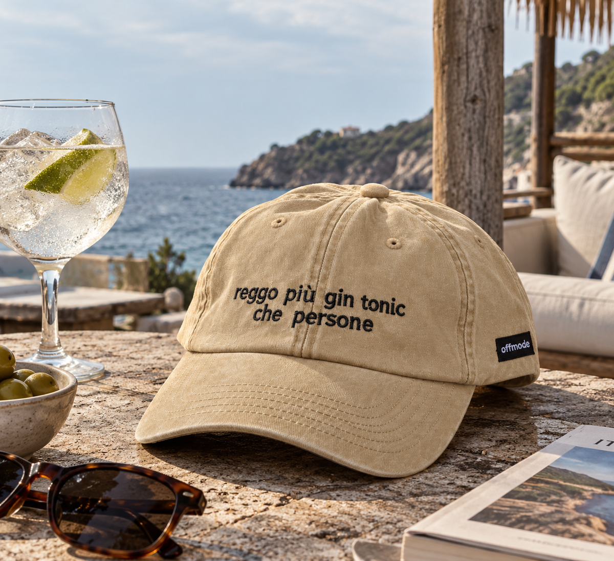 Cappello beige Offmode con la frase "reggo più gin tonic che persone" ricamata, accompagnato da un aperitivo con gin tonic al mare